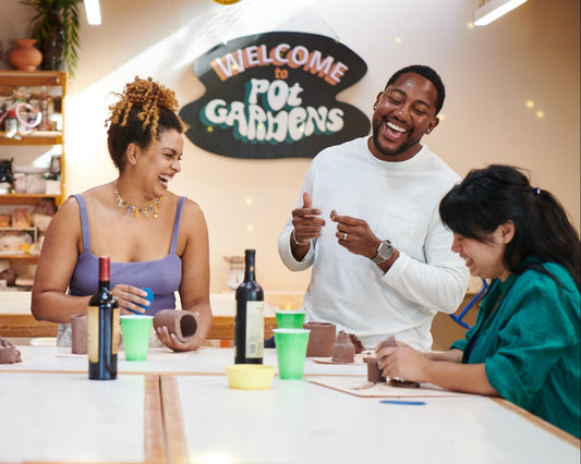 Group of people sitting around a table with drinks in a casual setting, with 'Welcome to Pot Gardens' sign in the background.